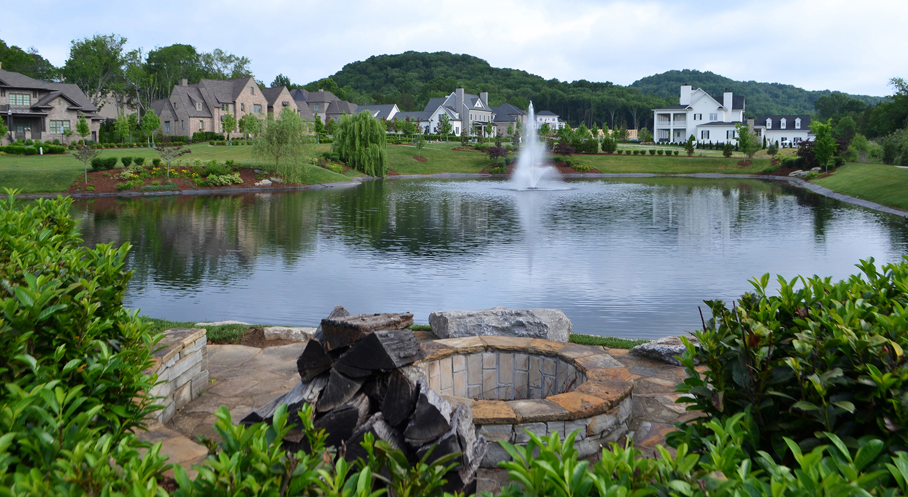 A stone fire pit overlooks a community pond with a fountain, surrounded by landscaped grounds and upscale homes with wooded hills in the background.
